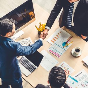 Businesspeople shaking hands on an office table piled high with documents and a laptop.